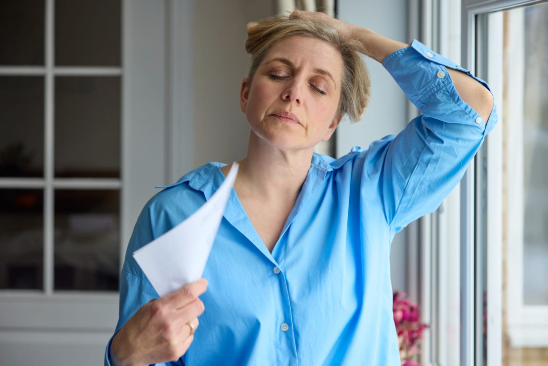 A woman with a headache is holding a tissue and standing by a window in her home.