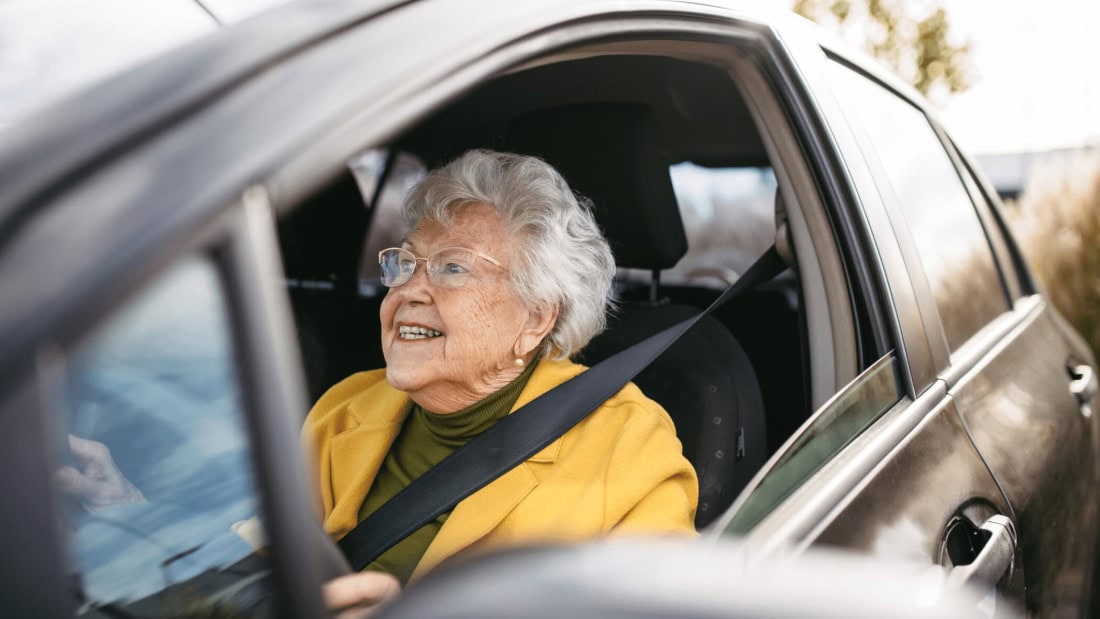 Oudere vrouw in auto met gordel, klaar voor rijbewijskeuring.