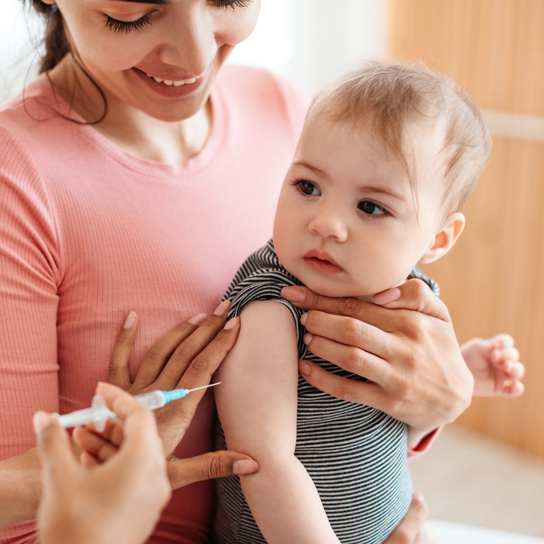 A baby is being vaccinated by a nurse, with the mother in the background.