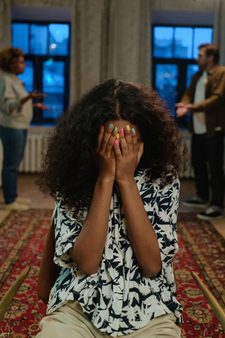 A woman with her hands over her face in a room with large windows and other people in the background.
